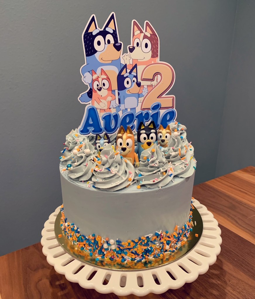Baby blue cake with buttercream swirls, Bluey cake topper and figurines, and blue and orange sprinkles